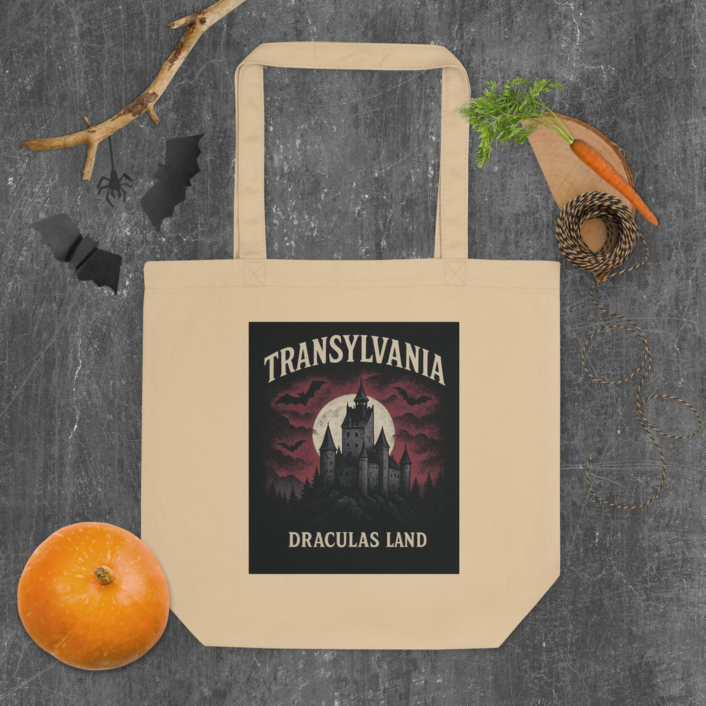 A beige canvas tote bag with a printed graphic of a gothic castle and the text 'TRANSYLIVANIA DRACULAS LAND' in a Halloween-themed layout, surrounded by autumn props like a small pumpkin, a carrot, a sprig of herbs, and black bat silhouettes on a dark textured background.