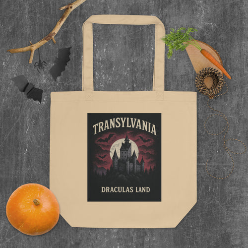 A beige canvas tote bag with a printed graphic of a gothic castle and the text 'TRANSYLIVANIA DRACULAS LAND' in a Halloween-themed layout, surrounded by autumn props like a small pumpkin, a carrot, a sprig of herbs, and black bat silhouettes on a dark textured background.