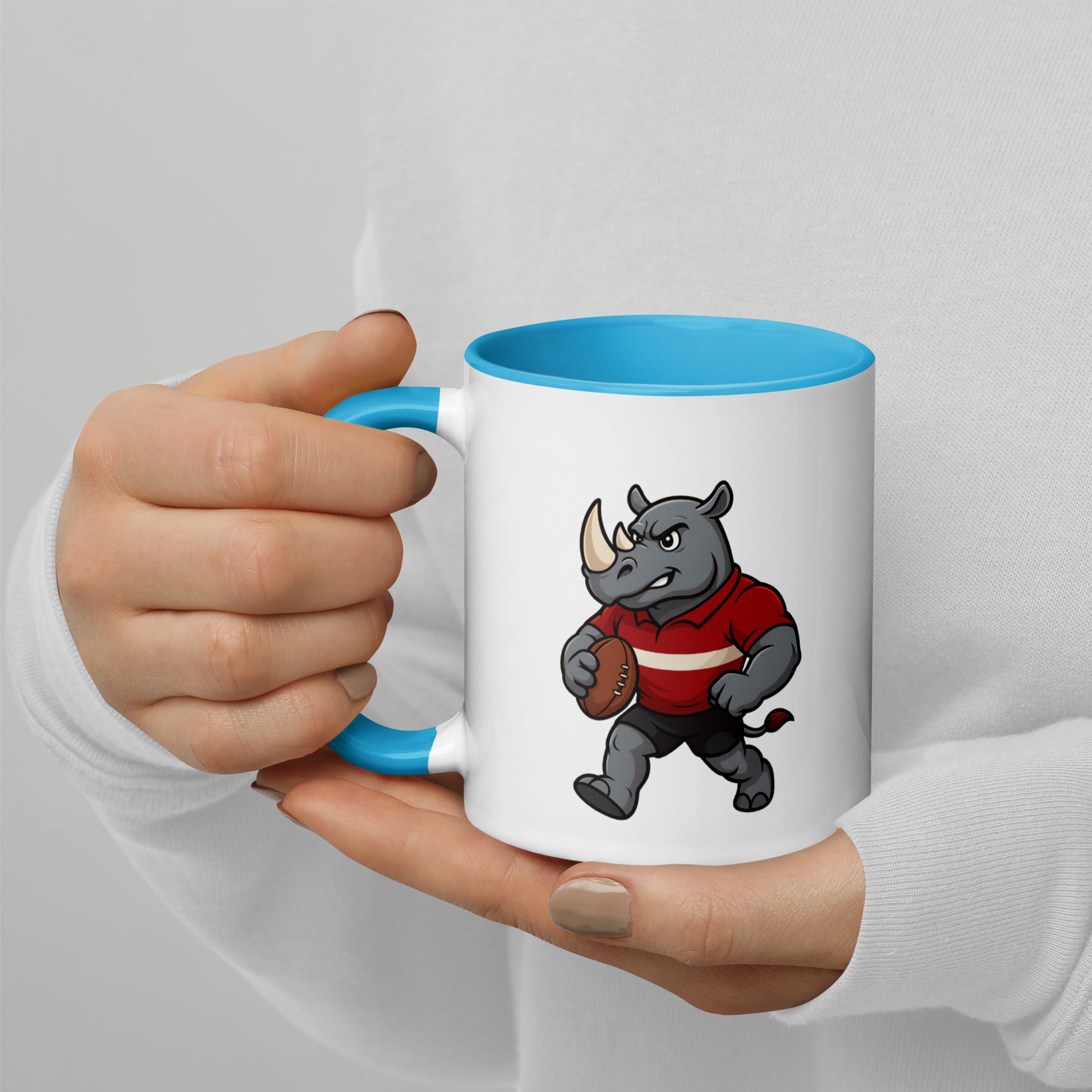 White mug with blue interior, featuring a rhino holding a rugby ball design, held by a person.
